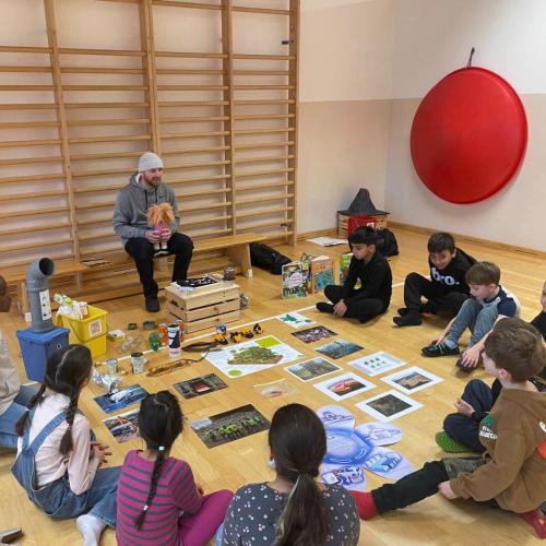 Sitzkreis mit Kindern, Robi und seiner Handpuppe Ronja, in der Mitte liegen Gegenstände und Bilder