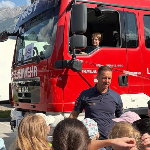 Feuerwehrmann spricht zu einer Kindergruppe, im Hintergrund sieht man einen Jungen, der im Feuerwehrauto sitzt