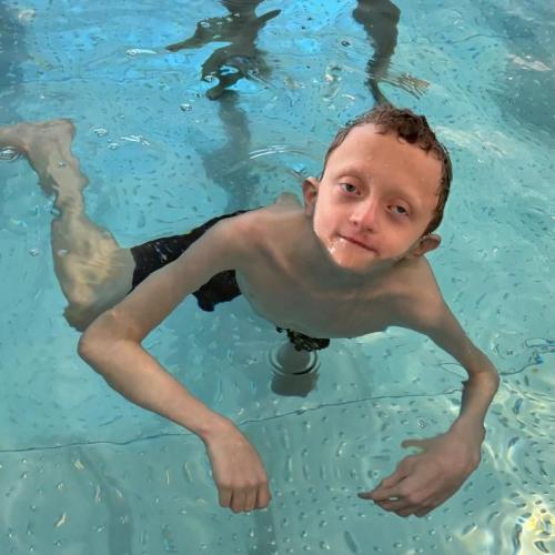 Bub gleitet durchs Wasser, im Hintergrund steht die Schwimmlehrerin mit einer Schwimmhilfe