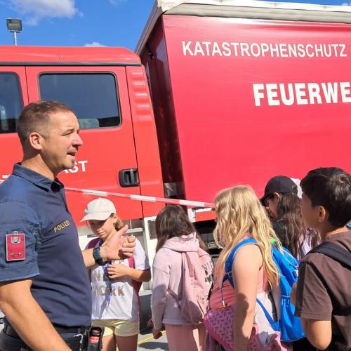 ein Polizist spricht mit den Kindern, im Hintergrund steht ein Feuerwehrauto